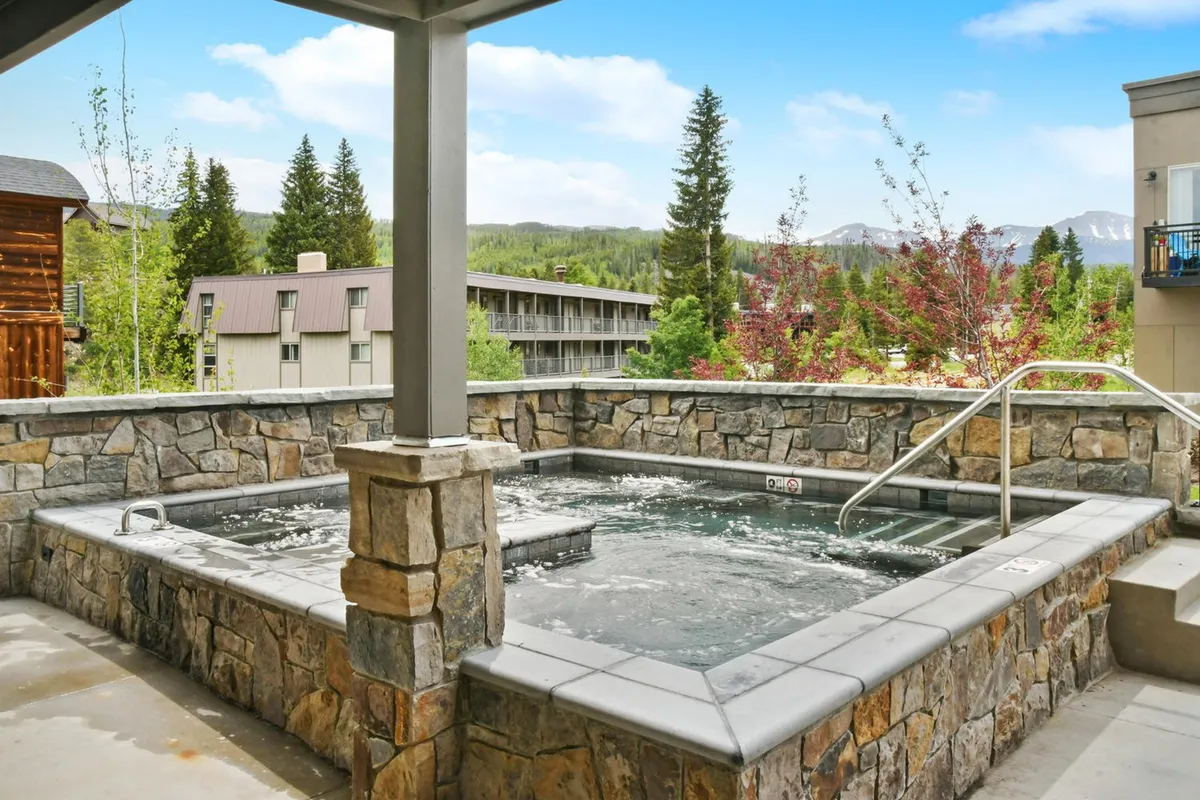 The hot tub area is featured here, set against a stunning mountain backdrop. The scene captures guests socializing and unwinding in a spacious, inviting hot tub while taking in sunset views and the natural landscape. The mood is tranquil and luxurious.