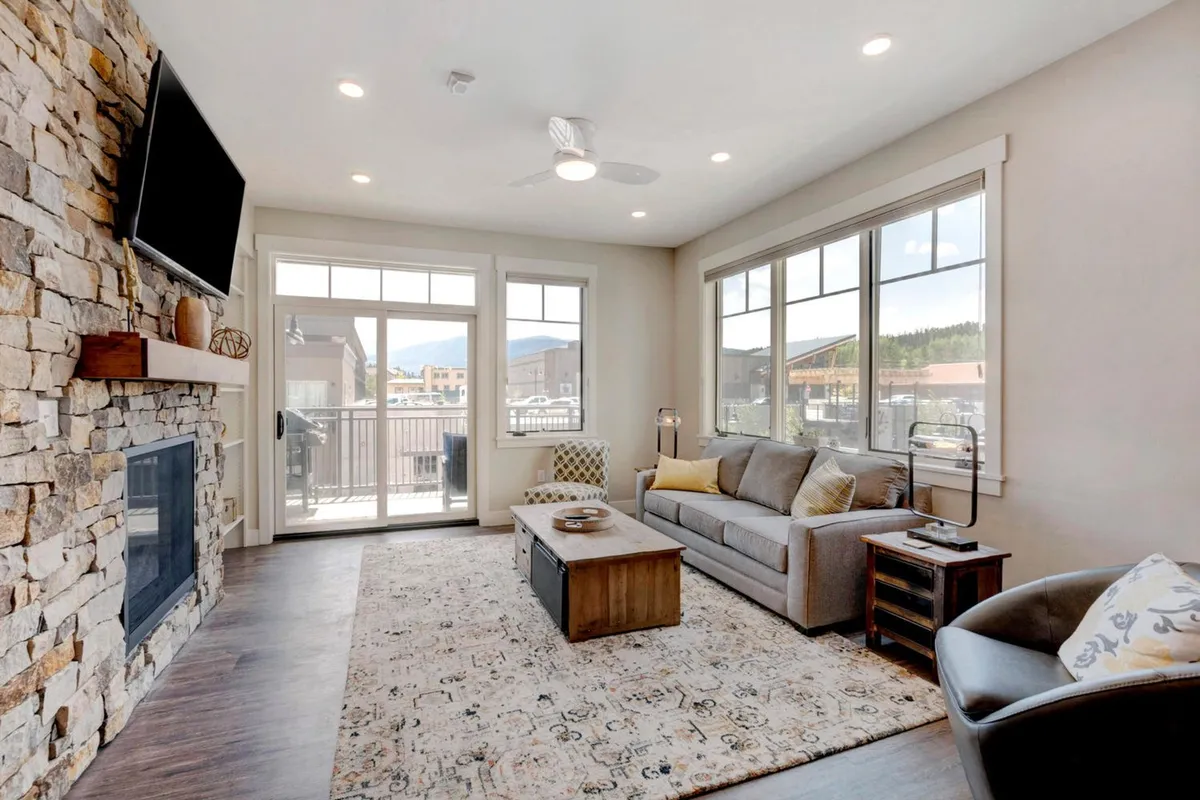 A cozy, open living space featuring a stone fireplace, large windows with beautiful outdoor views, a comfortable sofa, and a wooden coffee table. The room is set up for relaxation and socializing, offering a welcoming atmosphere perfect for gathering after a day of adventure.