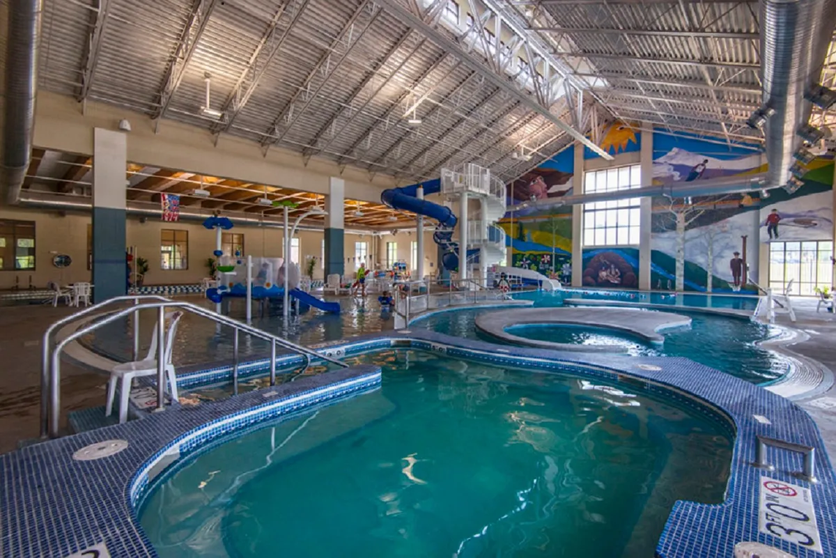 The interior of a recreation center's pool area features colorful murals, a winding water slide, a large swimming pool, and seating areas. Kids and families can be seen playing and swimming, creating a lively and energetic atmosphere ideal for young children.