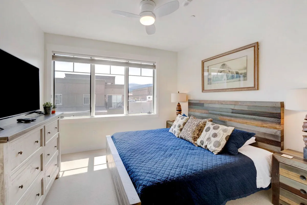 A bright, tranquil bedroom featuring a bed adorned with colorful pillows, large windows letting in gentle natural light, and a TV on the dresser. The room exudes comfort and calm, ideal for restful nights and peaceful mornings.