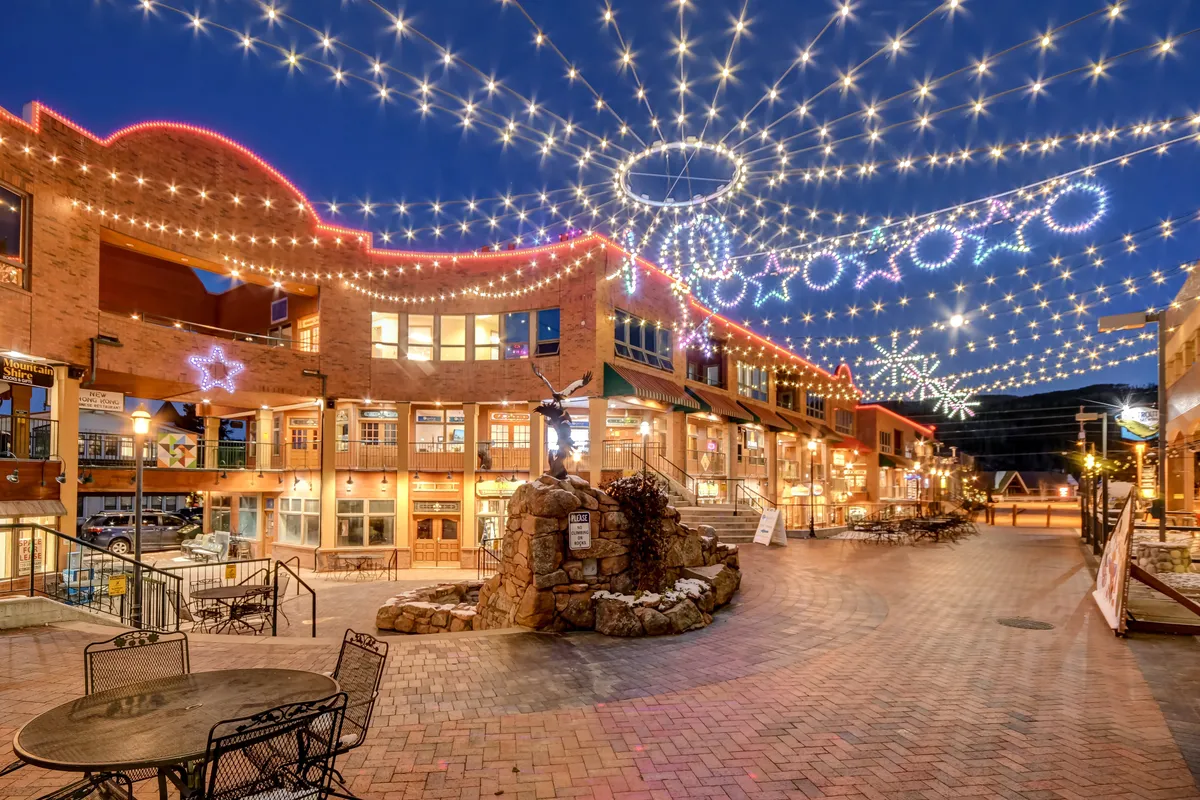 An inviting exterior scene featuring decorative string lights, a brick pathway, outdoor seating, a water feature, and surrounding building facades. The space is animated by people dining, shopping, socializing, and enjoying live music, with a touch of festive ambiance.