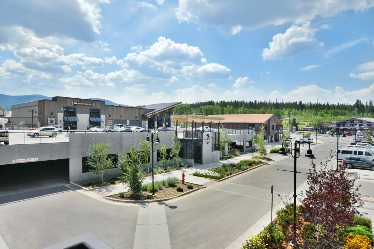 This exterior shot captures modern buildings set amongst landscaped areas, an open parking area, and a backdrop of trees and greenery. The lively atmosphere is evident, with people engaging in shopping, dining, walking, and socializing, which suggests a vibrant community environment.