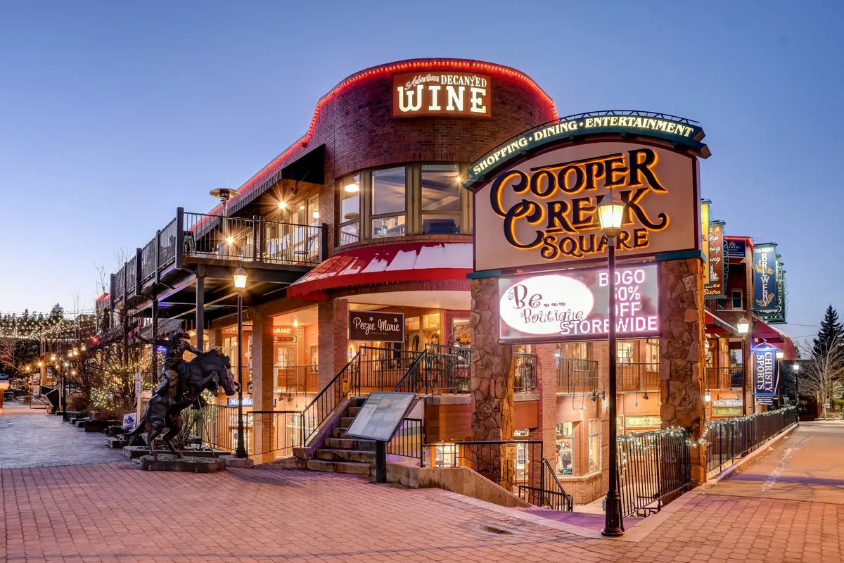 A lively exterior shot of Cooper Creek Square in downtown Winter Park, featuring inviting signs for shops and wine bars, an outdoor dining area, and artful touches like a horse statue. The atmosphere is bustling and festive, with clear indications of events and local culture.